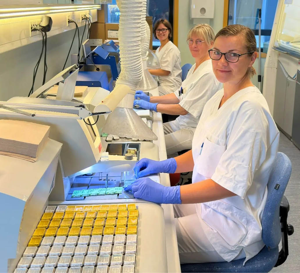 Personer i hvite arbeidskl r og bl hansker sitter ved en arbeidsbenk i et laboratorium og h ndterer sm  pr ver eller komponenter.  KI-generert beskrivelse