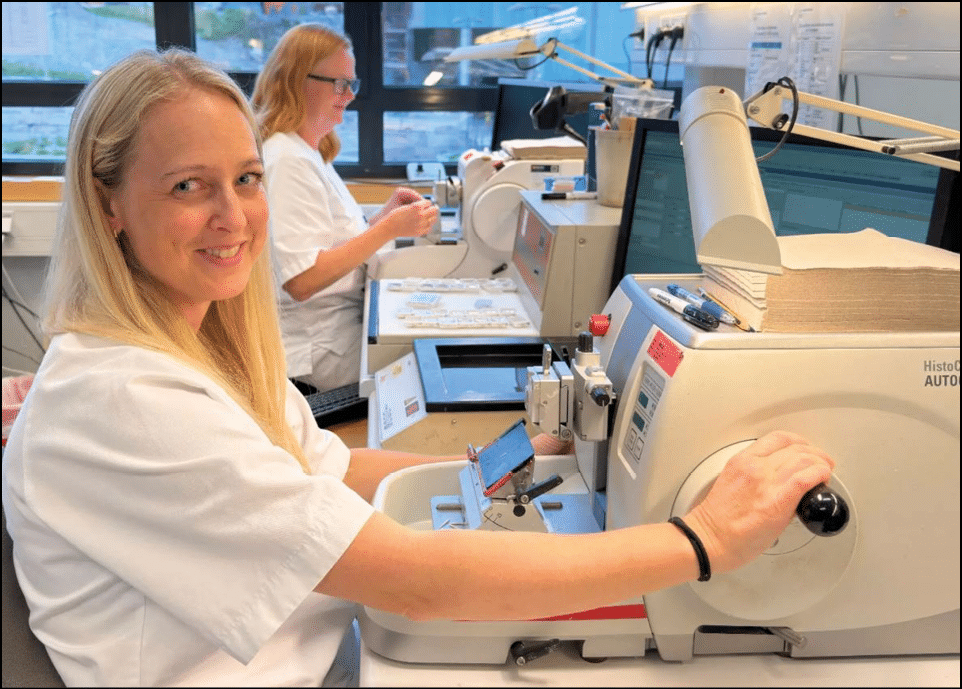 To personer med langt lyst h r og hvite kl r arbeider med medisinsk utstyr i et laboratorium. KI-generert beskrivelse