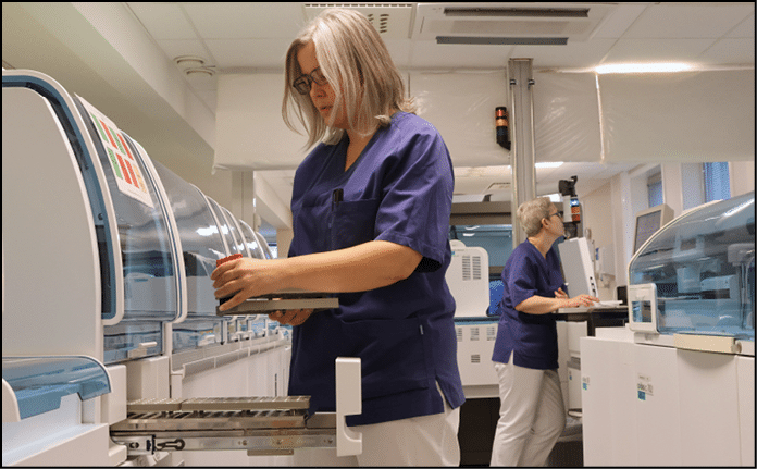 To personer i bl uniformer jobber med medisinsk laboratorieutstyr i et lyst rom.  KI-generert beskrivelse