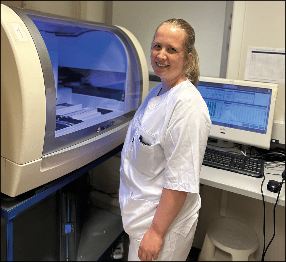 En person i hvit arbeidsuniform smiler og st r ved siden av en stor medisinsk eller laboratoriemaskin med bl tt lys og en dataskjerm i et klinisk rom. KI-generert beskrivelse